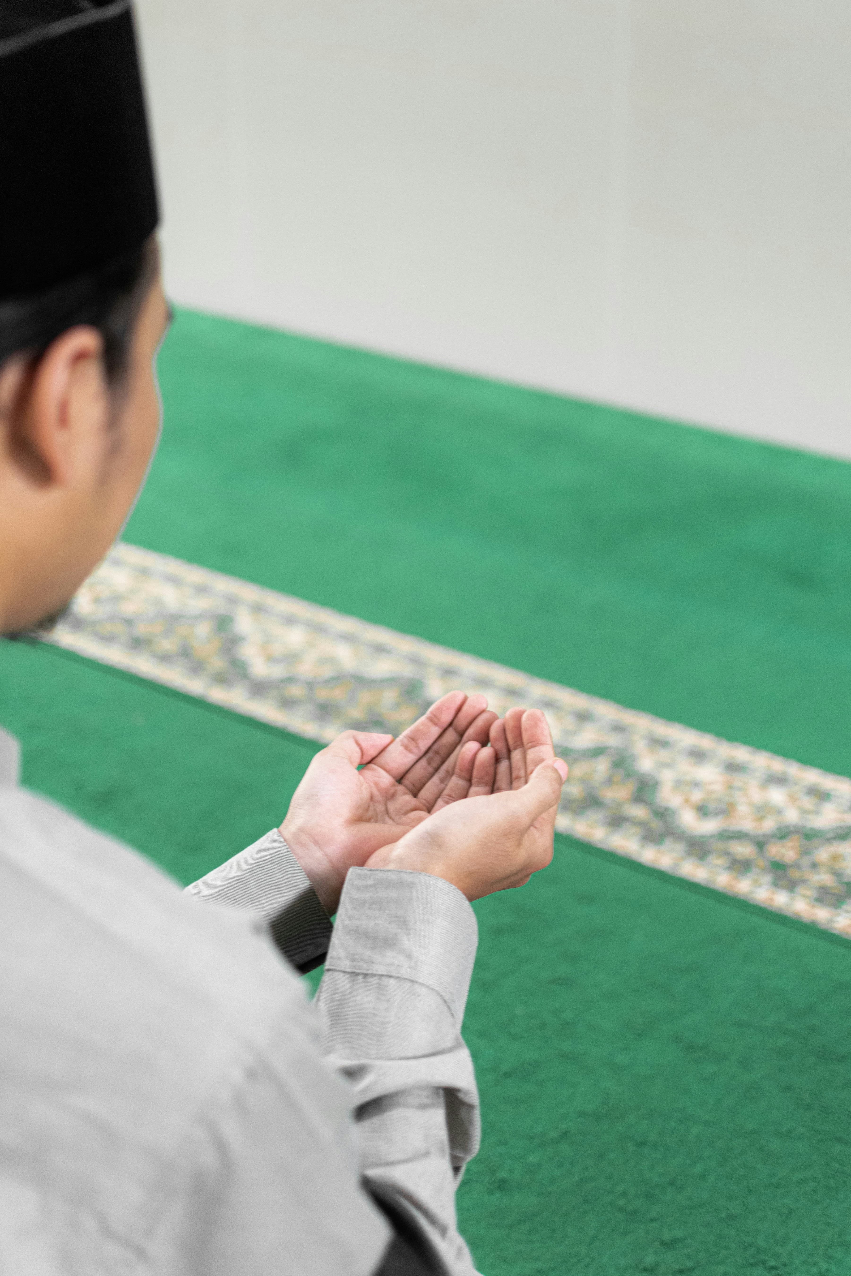 A person seen from behind making dua with palms raised, sitting on a green prayer rug