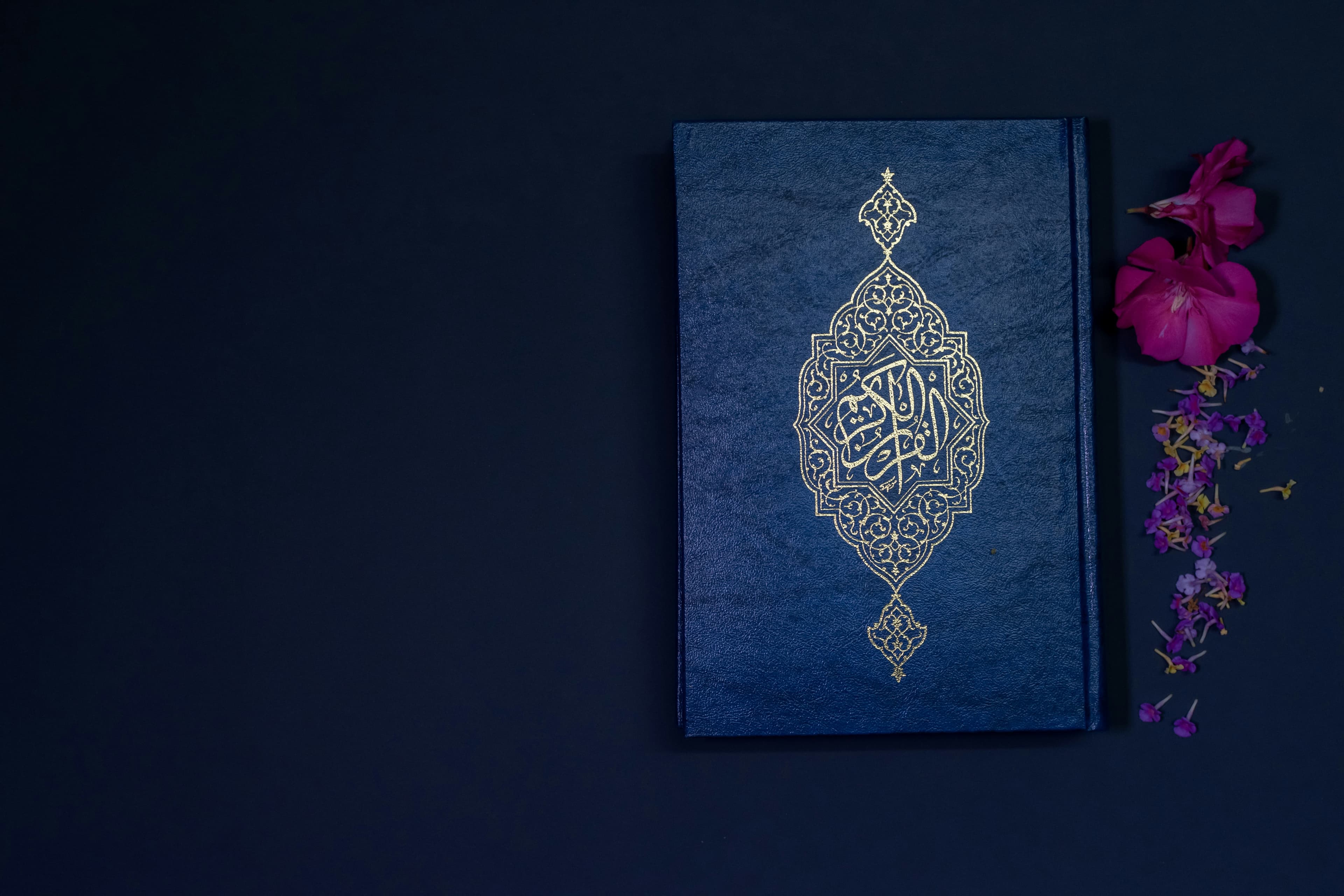 A closed navy Quran with gold calligraphy on the cover, beside pink flowers on a dark surface