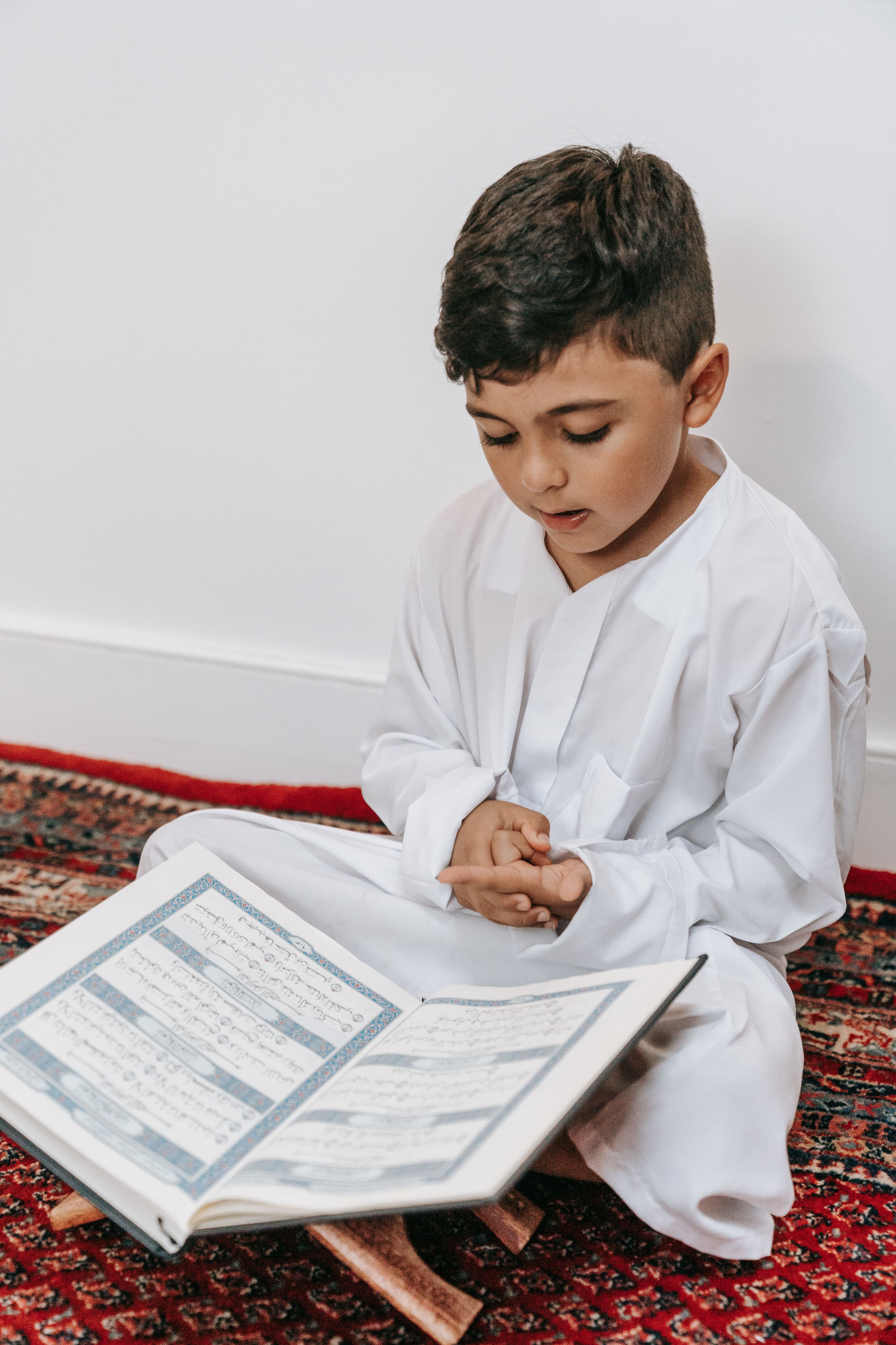 A young boy in a white thobe reading from the Quran on a wooden stand, seated on a prayer rug